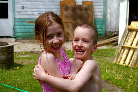 Girl and boy smiling and hugging while being sprayed by an outdoor sprinkler